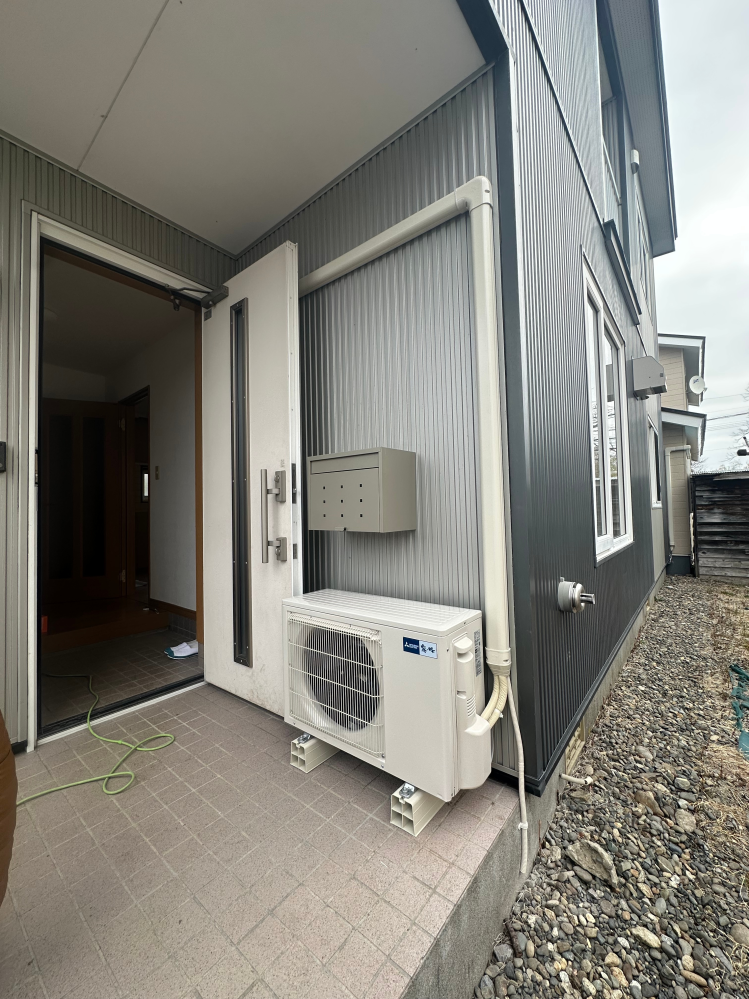 エアコンの室外機、設置場所について質問です。 中古住宅を購入しリフォーム中なのですが、室外機が写真のようにポストの真下、玄関ポーチ内に設置されていました。 屋根の形や落雪、雨だれを考慮してこの位置に設置したそうです。設置前に業者さんからは相談など一切なく現場監督さんに相談すると知らなかったの⁈と驚かれました。 とりあえず設置場所の変更は対応してもらえるのでしたいのですが、 ①ポーチ内のまま90度回転させて風向きを外に向け、カバーなどで目隠しをする ②南側の灯油ストーブの排気口の上部に来るよう高台を設置するか悩んでいます。 このままだと玄関扉を開けるとギリギリ、小さい子がいるので熱風が顔近くにかかる、視界に入る位置にあるので好奇心から指を入れそうなこと、来客の際に熱風がかかることもありこのままの状態だけは避けたいです。 エアコンは冷房のみで、猛暑日くらいしか使いません。（北海道の道東） 使用頻度は少ないですが、唯一道路に面して見えるとこで見栄えも気になります。 壁掛けは振動とかもあるので避けた方がいいのかなと思い、上記の二択で考えてます。 他にアドバイス等あれば教えていただきたいです。よろしくお願いします。